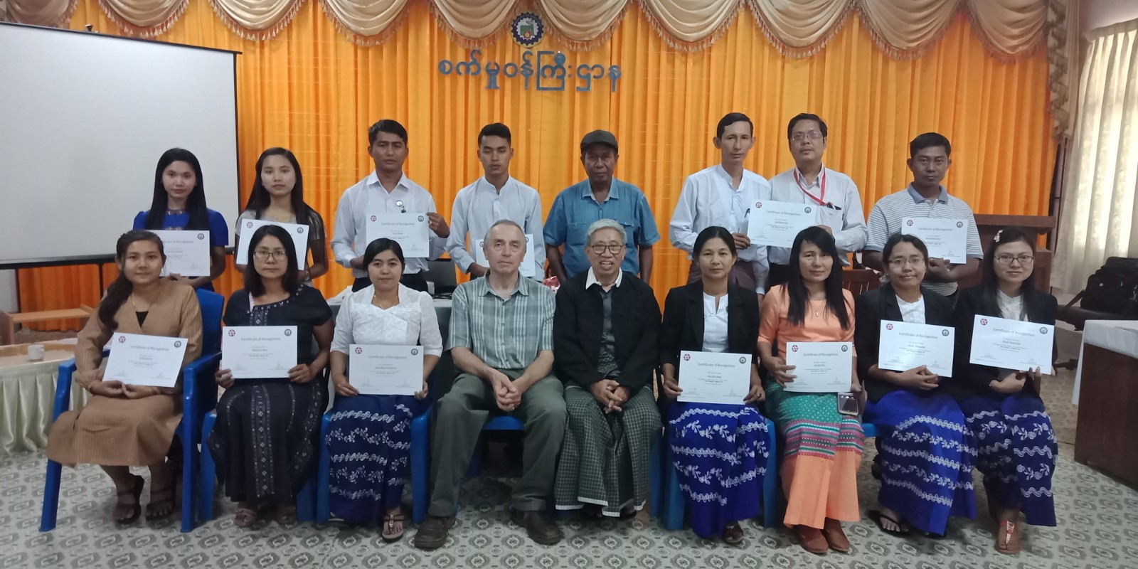 17 people in Myanmar sit in rows in front of a stage holding certificates